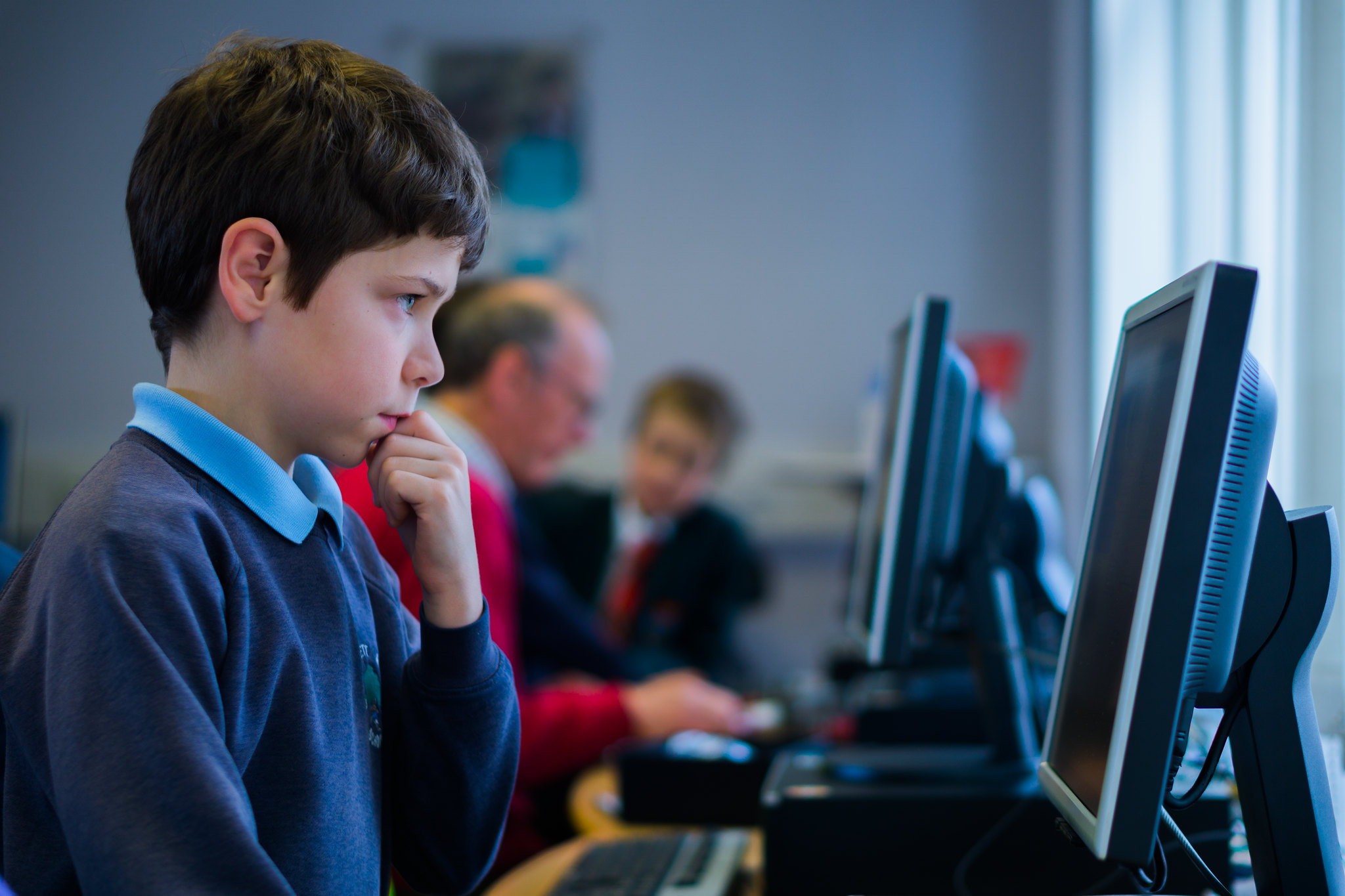 A row of computers being used, with a young boy using one in the foreground and people of other ages using computers in the background.