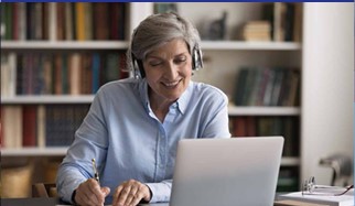 A woman wearing headphones takes notes as she watches something on a laptop computer.