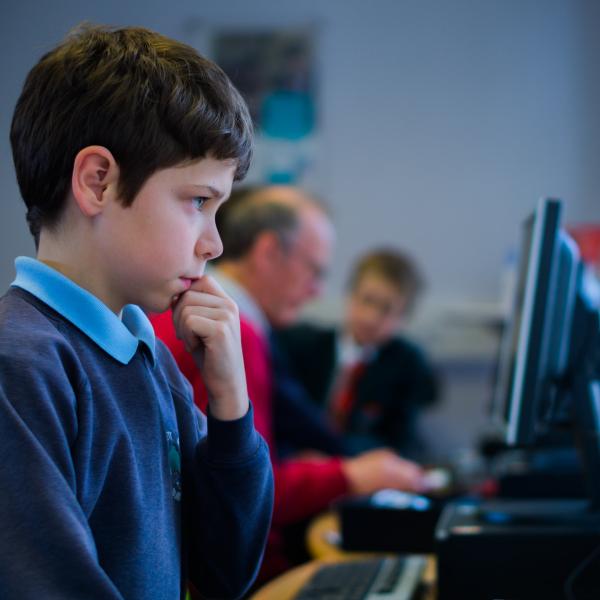 A row of computers being used, with a young boy using one in the foreground and people of other ages using computers in the background.